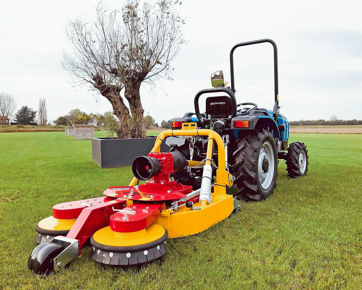 Blauwe compacttractor met Ilmer KMG cirkelmaaier op een gazon naast een knotwilg