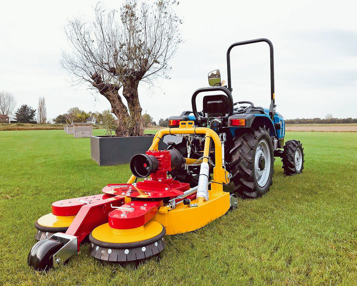 Blauwe compacttractor met Ilmer KMG cirkelmaaier op een gazon naast een knotwilg