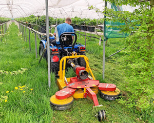 Ilmer KMG cirkelmaaier met twee zwenkschijven achter een blauwe compacttractor, maaiend tussen de stellingen in een tunnelkas