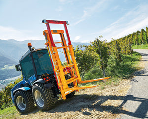 Blauwe smalspoortractor met Ilmer K2-D hefmast en vorken op de weg langs de wijngaard